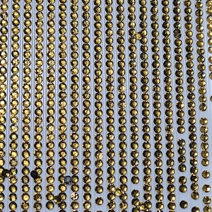 May include: A sheet of gold-colored rhinestones arranged in rows on a white background. The rhinestones are small and round, and they have a shiny, sparkly finish.