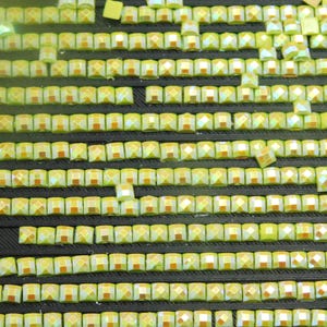 May include: Close-up of a collection of small, square, iridescent yellow rhinestones. The rhinestones are arranged in neat rows on a black surface, creating a textured pattern. The gems have a faceted design, reflecting light.