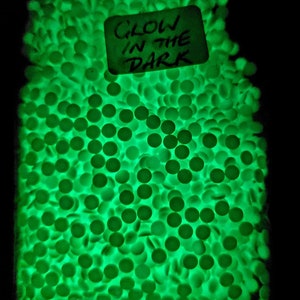 May include: A close-up image of a large quantity of small, round, green glow-in-the-dark beads. A white card with black writing reads "GLOW IN THE DARK".