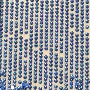 May include: Rows of iridescent blue rhinestones are arranged in a grid pattern on a white surface. A pile of loose rhinestones is in the bottom of the image.