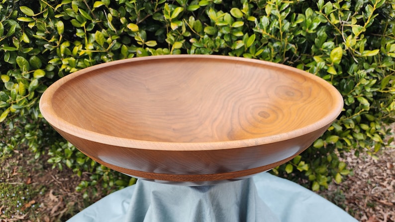 May include: A large, wooden bowl with a natural wood finish. The bowl is made of light brown wood with a grain pattern. The bowl is sitting on a blue cloth.