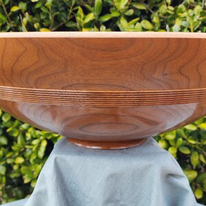May include: A large, wooden bowl with a natural wood finish. The bowl has a wide, rounded base and a deep, circular interior. The bowl is made of a light brown wood with a smooth, polished surface. The bowl has a decorative band of wood around the top edge.