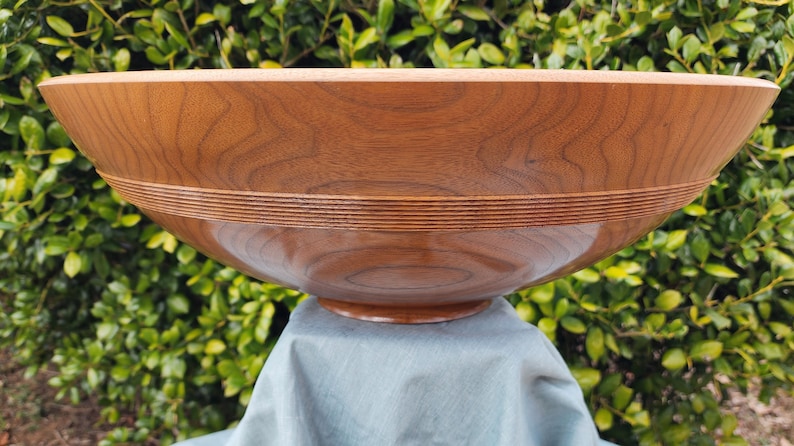 May include: A large, wooden bowl with a natural wood finish. The bowl has a wide, rounded base and a deep, circular interior. The bowl is made of a light brown wood with a smooth, polished surface. The bowl has a decorative band of wood around the top edge.