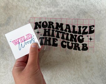 May include: A black vinyl sticker with the text "Normalize hitting the curb" in a retro font. The sticker is on a clear sheet with a grid pattern.
