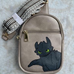 Könnte beinhalten: Eine beige Kunstleder-Umhängetasche mit einem schwarzen und grünen Drachen-Motiv. Die Tasche hat einen braunen und weißen gemusterten Riemen.