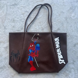 May include: A dark brown tote bag featuring a graphic of Spider-Man in a crouching pose, a black spider emblem on the left side, and the text SPIDER-MAN in white block letters on the right side. The bag has two thin shoulder straps and a gold snap.