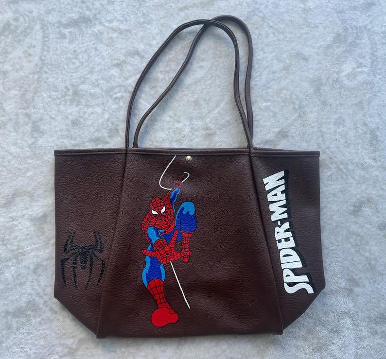May include: A dark brown tote bag featuring a graphic of Spider-Man in a crouching pose, a black spider emblem on the left side, and the text SPIDER-MAN in white block letters on the right side. The bag has two thin shoulder straps and a gold snap.