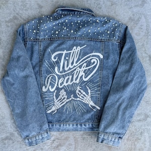 Puede incluir: Una chaqueta vaquera azul con adornos de perlas en los hombros y un gráfico blanco en la espalda que dice "Till Death" con dos manos de esqueleto haciendo una promesa meñique.