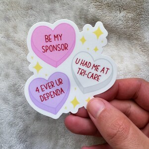May include: A sticker featuring three heart-shaped candies with text. The pink heart says "BE MY SPONSOR," the gray heart says "U HAD ME AT TRI-CARE," and the purple heart says "4 EVER UR DEPENDA." Yellow star accents are also included.