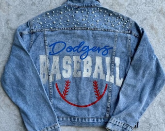May include: A light blue denim jacket with a white "Dodgers Baseball" graphic and a baseball design on the back. The jacket is decorated with pearl-like embellishments on the shoulders and collar.