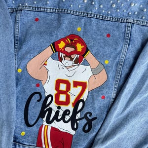 May include: A light blue denim jacket with a white, red, and yellow illustration of a football player wearing a jersey with the number 87 and the word "Chiefs". The jacket is decorated with white pearl-like embellishments.