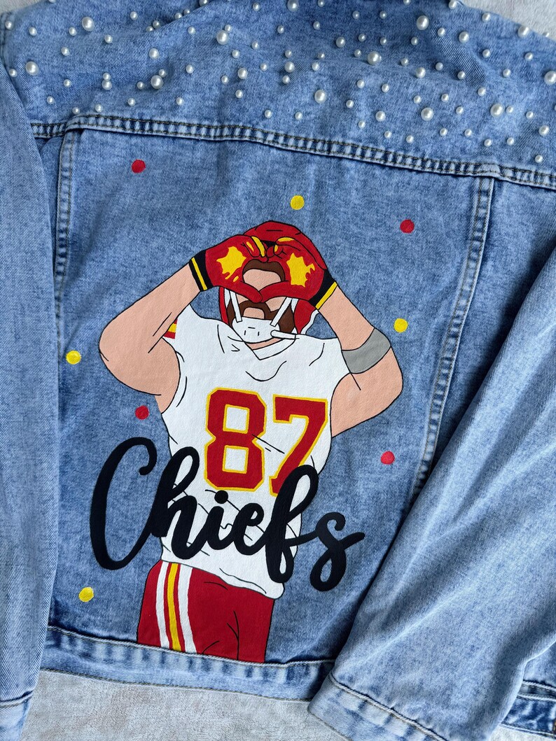 May include: A light blue denim jacket with a white, red, and yellow illustration of a football player wearing a jersey with the number 87 and the word "Chiefs". The jacket is decorated with white pearl-like embellishments.