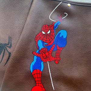 May include: A brown textured tote bag featuring a graphic illustration of the superhero Spider-Man in his iconic red and blue suit. The character is depicted in a dynamic pose with a white web line extending upwards against the dark brown leather-like material.