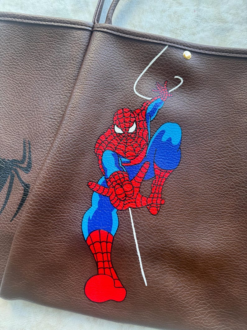 May include: A brown textured tote bag featuring a graphic illustration of the superhero Spider-Man in his iconic red and blue suit. The character is depicted in a dynamic pose with a white web line extending upwards against the dark brown leather-like material.