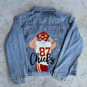 May include: A blue denim jacket with a white pearl embellishment on the back. The back of the jacket features a red, yellow, and white illustration of a football player with the number 87 and the word "Chiefs" in black.