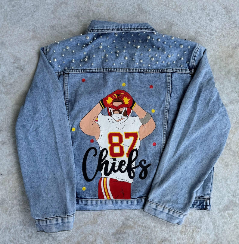 May include: A blue denim jacket with a white pearl embellishment on the back. The back of the jacket features a red, yellow, and white illustration of a football player with the number 87 and the word "Chiefs" in black.