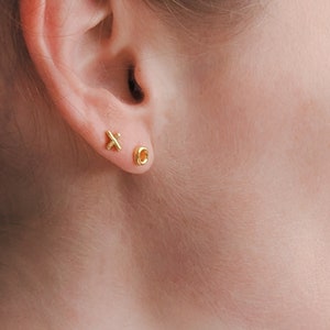 May include: A close-up of a person's ear with two gold earrings. The left earring is a small, simple circle. The right earring is a small, simple 'x'.