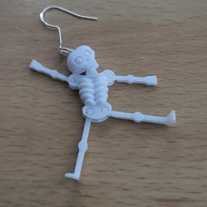 May include: White skeleton earring with a silver hook. The skeleton is posed with its arms outstretched and one leg raised.
