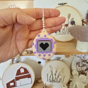 May include: A beige and purple pendant with a pixelated black heart design, on a white beaded chain. The pendant is surrounded by embroidered hoops with floral and barn designs, and a felt mushroom.