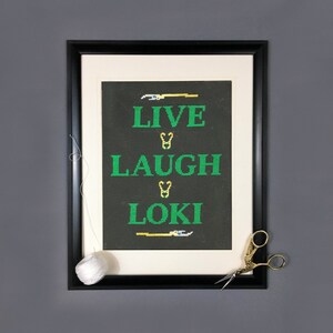 Può includere: Un ricamo a punto croce incorniciato con sfondo nero e testo verde che recita "Live Laugh Loki". Il testo è separato da elementi decorativi dorati. Un gomitolo di filo bianco e un paio di forbici dorate sono sul tavolo sotto la cornice.