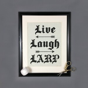 May include: Black framed embroidery with white background and black text that reads "Live Laugh LARP" with arrows between each word.