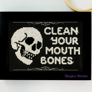Könnte beinhalten: Schwarz-weiß gestickte Kreuzstich-Stickerei in einem schwarzen Rahmen. Die Stickerei zeigt einen Schädel mit dem Text "Clean Your Mouth Bones" und der Signatur "Sleepless Stitcher".