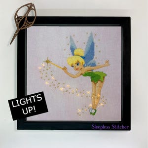 Könnte beinhalten: Ein gerahmtes Kreuzstichbild von Tinkerbell, der Disney-Fee, mit einem Zauberstab und funkelnden Lichtern. Der Rahmen ist schwarz und der Kreuzstich ist auf einem hellvioletten Hintergrund. Der Text "Lights Up!" befindet sich in der linken unteren Ecke des Rahmens. Der Text "Sleepless Stitcher" befindet sich in der rechten unteren Ecke des Rahmens.