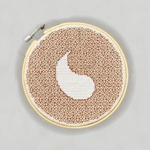 May include: A white and brown embroidery hoop with a geometric pattern in brown thread. The pattern is a yin yang symbol with a white background.