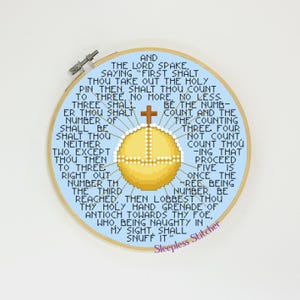 Op de afbeelding: Een blauwe en witte borduurring met een kruis en een gele cirkel in het midden. De ring heeft tekst rond de rand die luidt: "En de Heer sprak, en zei: "Eerst zul je de heilige pin eruit halen, dan zul je tot drie tellen, niet meer, niet minder. Drie zal het aantal zijn dat je moet tellen, en het tellen drie vier niet tellen tellen je -ing dat doorgaan vijf is een keer de -ree, zijnde nummer zijn bereikt, dan gooi je je heilige handgranaat van Antiochië naar je vijand, die, omdat hij stout is in mijn zicht, het moet doven." De tekst is in wit. De ring bevindt zich op een lichtblauwe achtergrond.