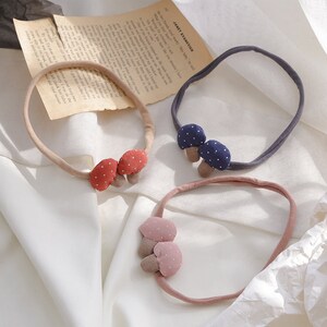 May include: Three fabric headbands with mushroom-shaped embellishments. The headbands are in navy blue, pink, and red. The mushrooms have white polka dots.