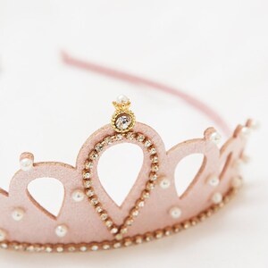 May include: A pink felt crown with a gold center piece and pearl accents. The crown is decorated with rhinestones.