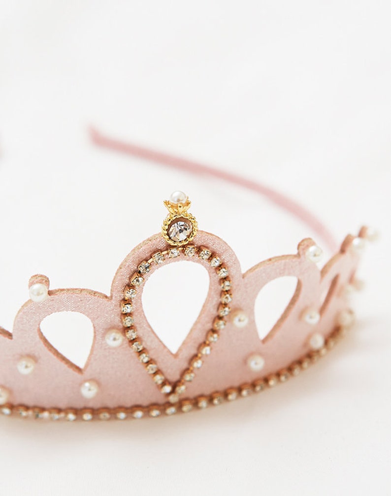 May include: A pink felt crown with a gold center piece and pearl accents. The crown is decorated with rhinestones.