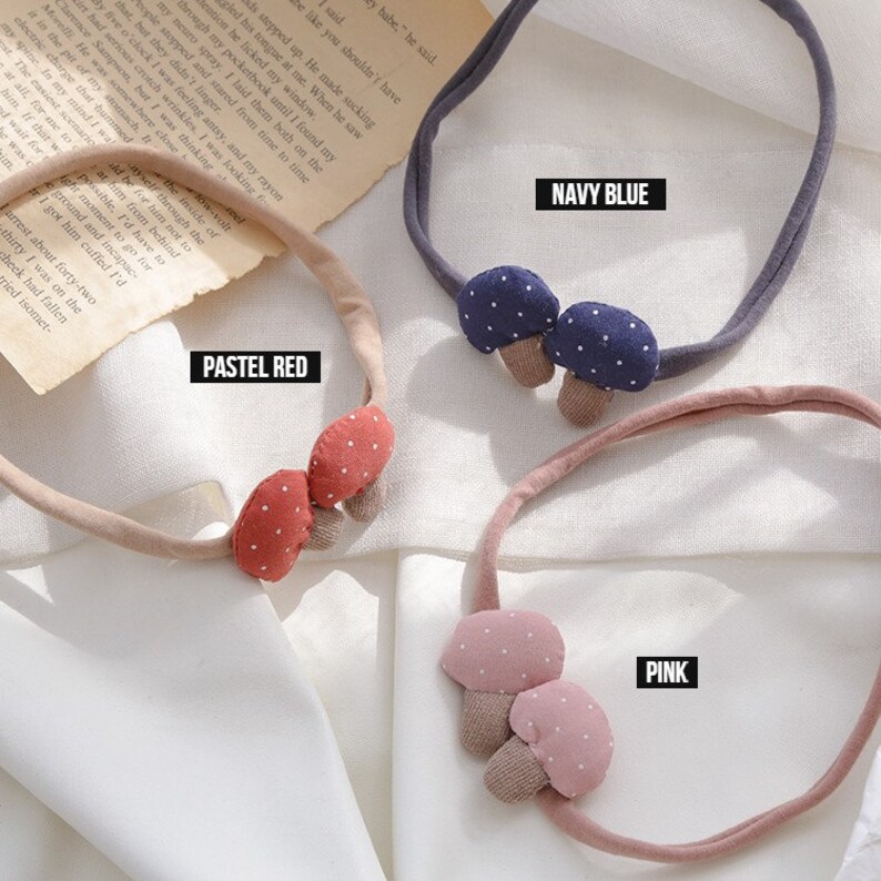 May include: Three headbands with mushroom-shaped embellishments. The headband on the left has red mushrooms with white polka dots, the middle headband has navy blue mushrooms with white polka dots, and the headband on the right has pink mushrooms with white polka dots. The headbands are on a white surface. The text "PASTEL RED", "NAVY BLUE", and "PINK" are written below each headband.