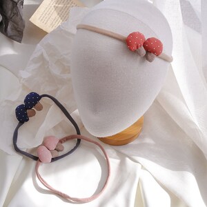 May include: Two headbands with mushroom-shaped fabric embellishments. One headband is pink with white polka dots and the other is navy blue with white polka dots.