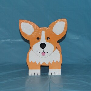 May include: A wooden figurine of a corgi dog with orange and white paint. The dog has a black nose and a pink tongue.