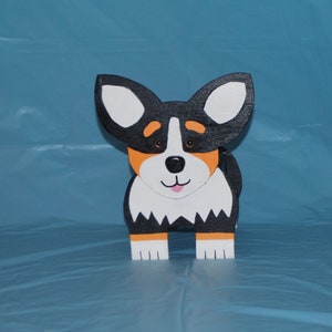 May include: A wooden figurine of a black, white, and orange corgi dog. The dog is standing on all four legs and has a happy expression on its face.