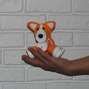 May include: A handcrafted orange and white corgi dog figurine. The dog has a black nose and button eyes. The figurine is made of ceramic or a similar material. The dog is sitting with its legs outstretched. The ears are white with orange trim.