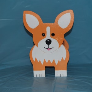 May include: A wooden toy dog shaped like a corgi. The dog is orange and white with black eyes and a pink tongue.