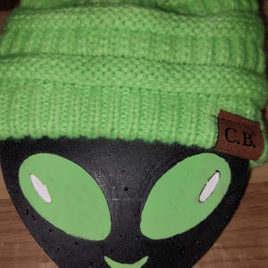 May include: A green knitted beanie with a brown leather patch that says "C.B." on it. The beanie is sitting on top of a black alien face with green eyes.