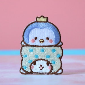 May include: Embroidered iron-on patch featuring a blue penguin wearing a yellow crown and a white polka dot blanket. The penguin is smiling and has a small, white, smiling penguin peeking out from under the blanket.