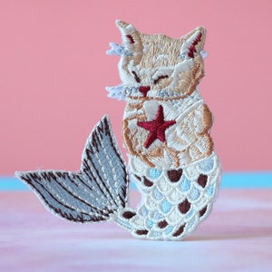 May include: Embroidered iron-on patch of a cat with a mermaid tail. The cat is light brown with a red star on its chest. The tail is blue and white with brown accents.