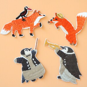 Puede incluir: Cuatro parches bordados para planchar que representan un zorro con un pájaro en la espalda, un zorro con un sombrero y dos pingüinos, uno tocando la trompeta y el otro sosteniendo una batuta de director.