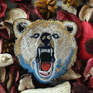 Könnte beinhalten: Ein gestickter Bären-Patch in Braun und Weiß mit einem blauen und weißen Rand. Der Bär hat seinen Mund weit geöffnet, wobei eine rote Zunge und schwarze Zähne zu sehen sind. Der Patch ist von getrockneten Blütenblättern umgeben.