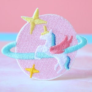 May include: A pink embroidered patch with a white unicorn with rainbow wings flying around a blue planet ring. The unicorn has a yellow star on its head and there are two yellow stars on the planet.