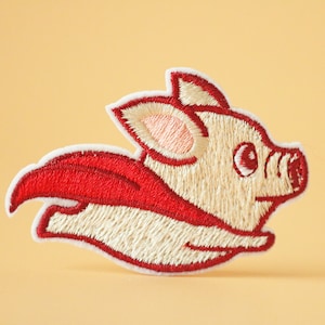 May include: Embroidered patch of a white pig wearing a red cape and flying. The pig has a pink ear and a black eye.