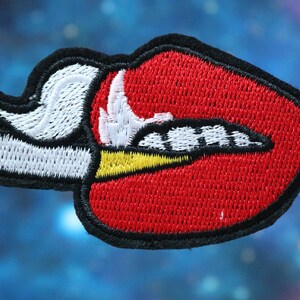 May include: Red embroidered lips with a white and yellow cigarette coming out of the side.
