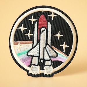 May include: A white and red embroidered space shuttle patch with a rainbow and stars on a black background. The patch is circular and has a white border.