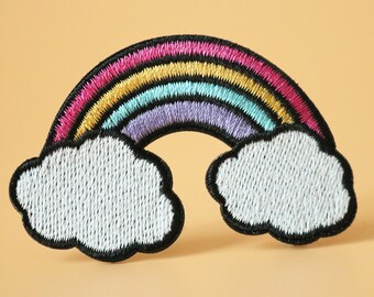Può includere: Una toppa ricamata con un arcobaleno colorato di rosa, giallo, blu e viola sopra due nuvole bianche. La toppa è bordata con filo nero.