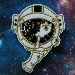 Puede incluir: Un parche bordado blanco y dorado de un casco de astronauta con una visera agrietada que revela un cielo estrellado negro y dorado.