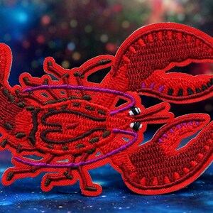 May include: Embroidered red lobster patch with purple and black detailing. The patch depicts a detailed lobster design with outstretched claws. The patch is likely for clothing or accessories.